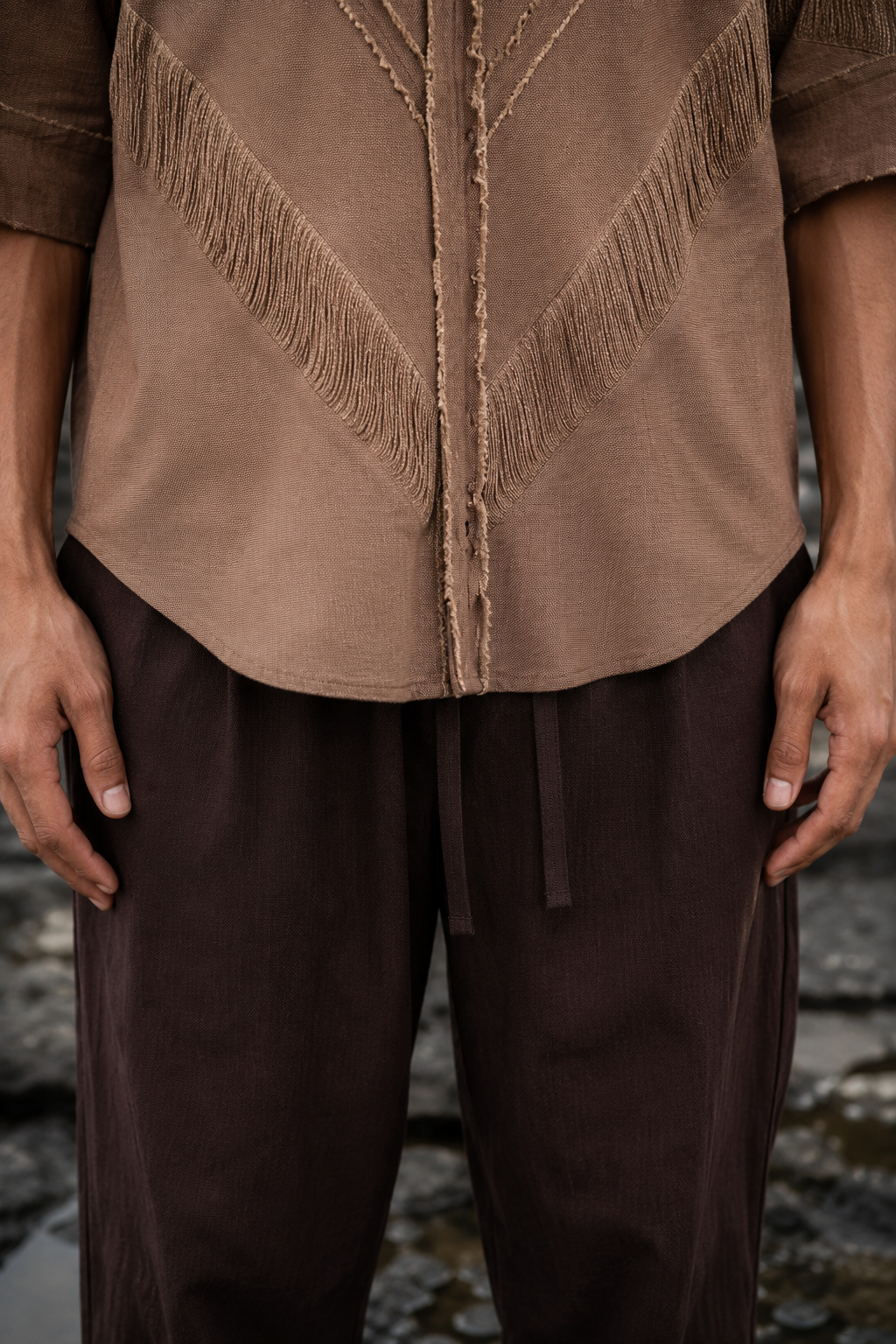 A close-up of a person wearing a brown textured top with fringe and drawstring details, paired with dark brown pants, set against a blurred outdoor background.