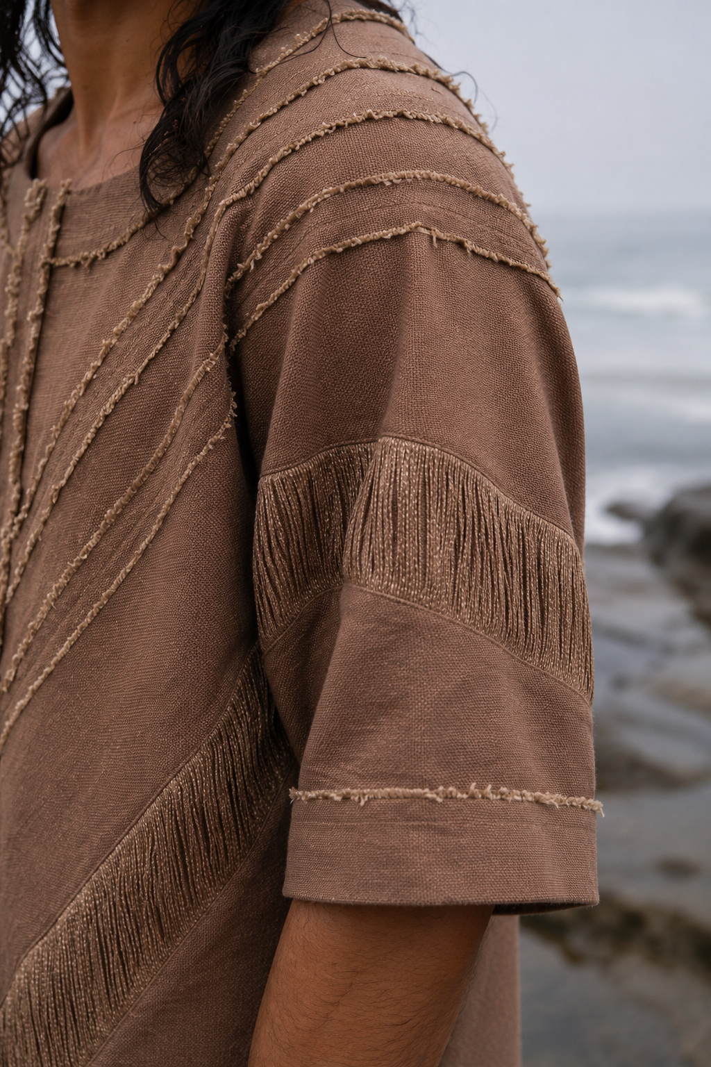 A close-up of a person wearing a brown textured short-sleeve top with fringe and stitched details, photographed outdoors near the ocean.