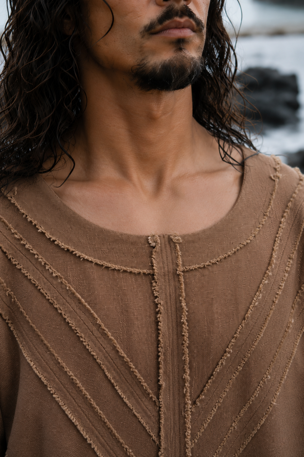 A close-up of a man wearing a brown textured knit top with a crew neck and vertical ribbed details, set against an outdoor blurred background.