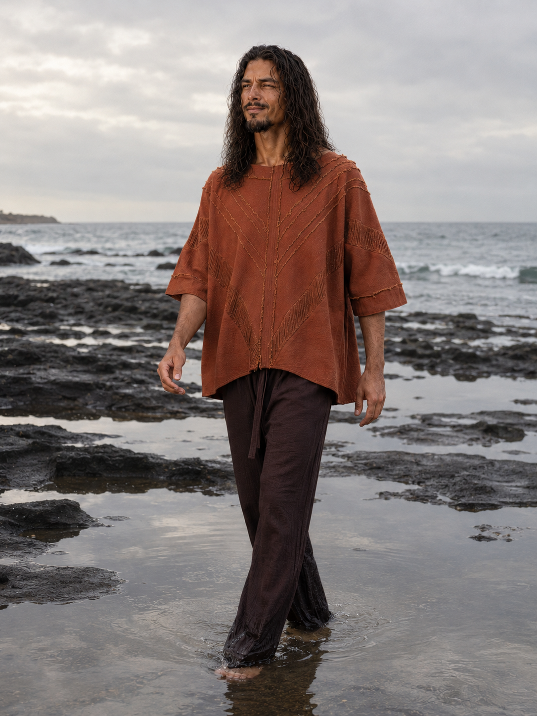 A man with long dark hair stands barefoot in shallow tide on a rocky beach, wearing a loose rust-orange textured top and dark brown pants.