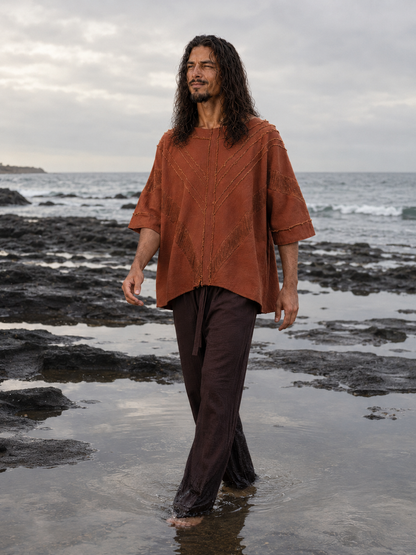 A man with long dark hair stands barefoot in shallow tide on a rocky beach, wearing a loose rust-orange textured top and dark brown pants.