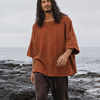 A man with long dark hair stands barefoot on a rocky ocean shore wearing a loose rust-orange short-sleeve top and dark brown pants.
