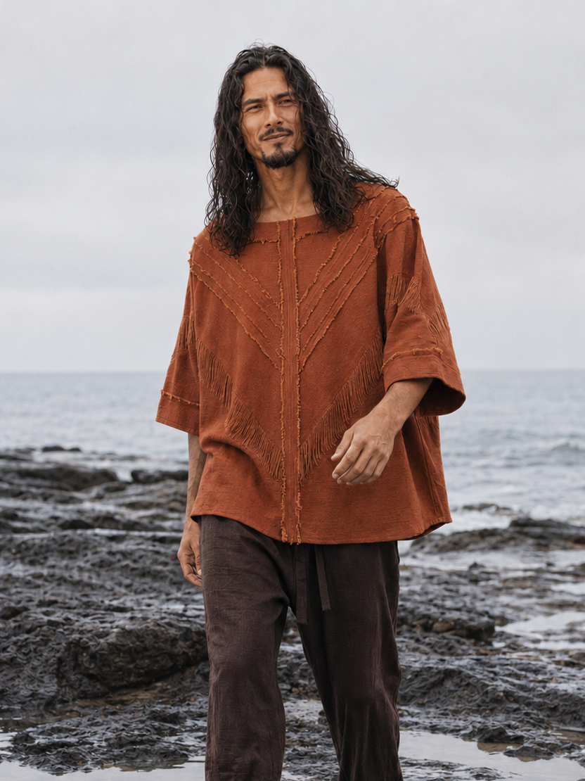 A man with long dark hair stands barefoot on a rocky ocean shore wearing a loose rust-orange short-sleeve top and dark brown pants.
