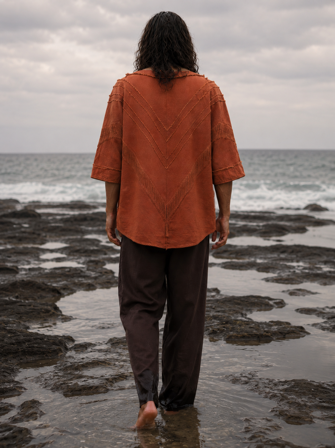 A person standing on a rocky beach facing the ocean, wearing a rust-orange short-sleeve button-up shirt with a textured chevron pattern on the back and dark brown pants.