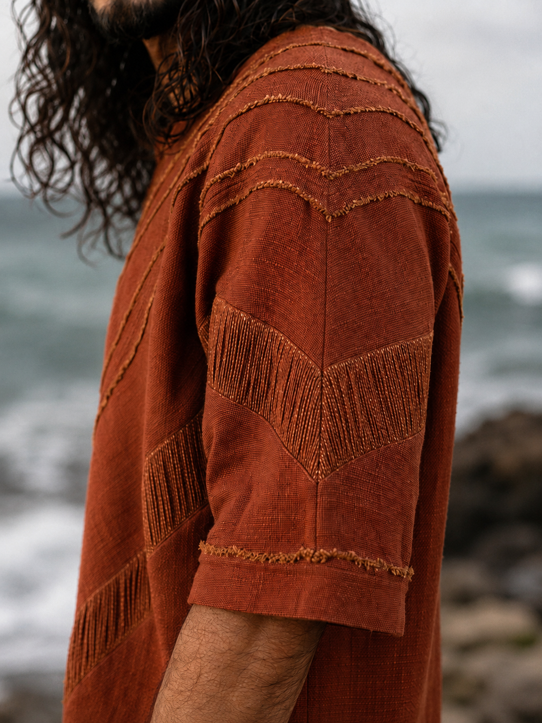 A close-up side view of a person wearing a rust-orange textured short-sleeve top with decorative fringe and zigzag stitched details, photographed outdoors near the ocean.