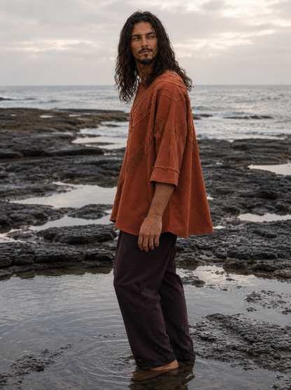 A man with long dark hair stands barefoot in shallow tide pools on a rocky ocean shore, wearing an oversized rust-orange short-sleeve top and dark brown pants.