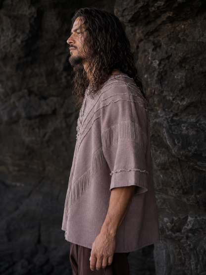 A man with long dark hair stands in profile against a dark rocky background, wearing a loose short-sleeve top in a muted dusty mauve or taupe color with fringe and textured trim details.