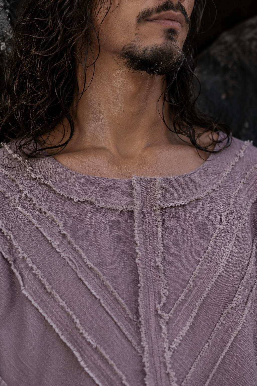 Close-up of a person wearing a textured mauve-lavender top with vertical and diagonal frayed ribbed details, shown against a dark natural background.
