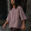 A man walking through shallow water at a rocky beach, wearing a loose dusty mauve linen shirt and dark brown pants.