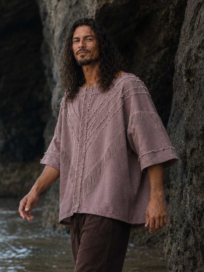 A man walking through shallow water at a rocky beach, wearing a loose dusty mauve linen shirt and dark brown pants.