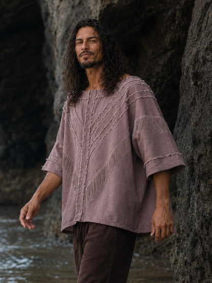 A man walking through shallow water at a rocky beach, wearing a loose dusty mauve linen shirt and dark brown pants.
