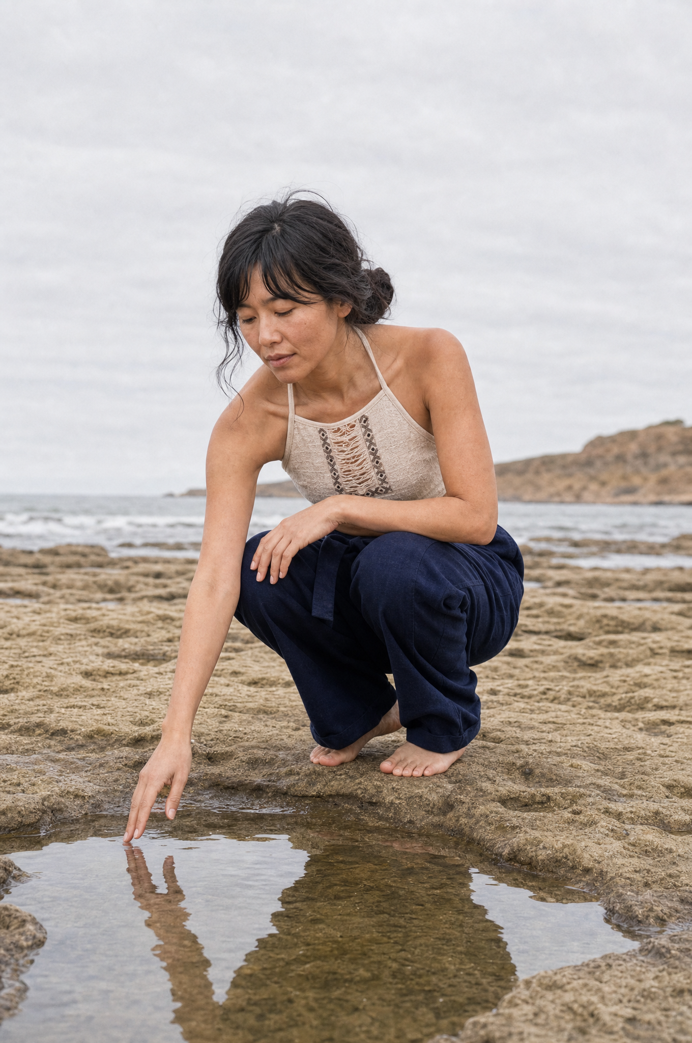 A woman crouches on a rocky beach next to a small tide pool, touching the water. She is wearing a beige crochet halter top and dark navy pants.