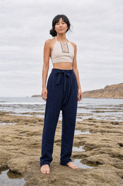 A woman stands barefoot on a rocky beach wearing navy blue high-waisted drawstring pants and a cream crochet halter crop top.