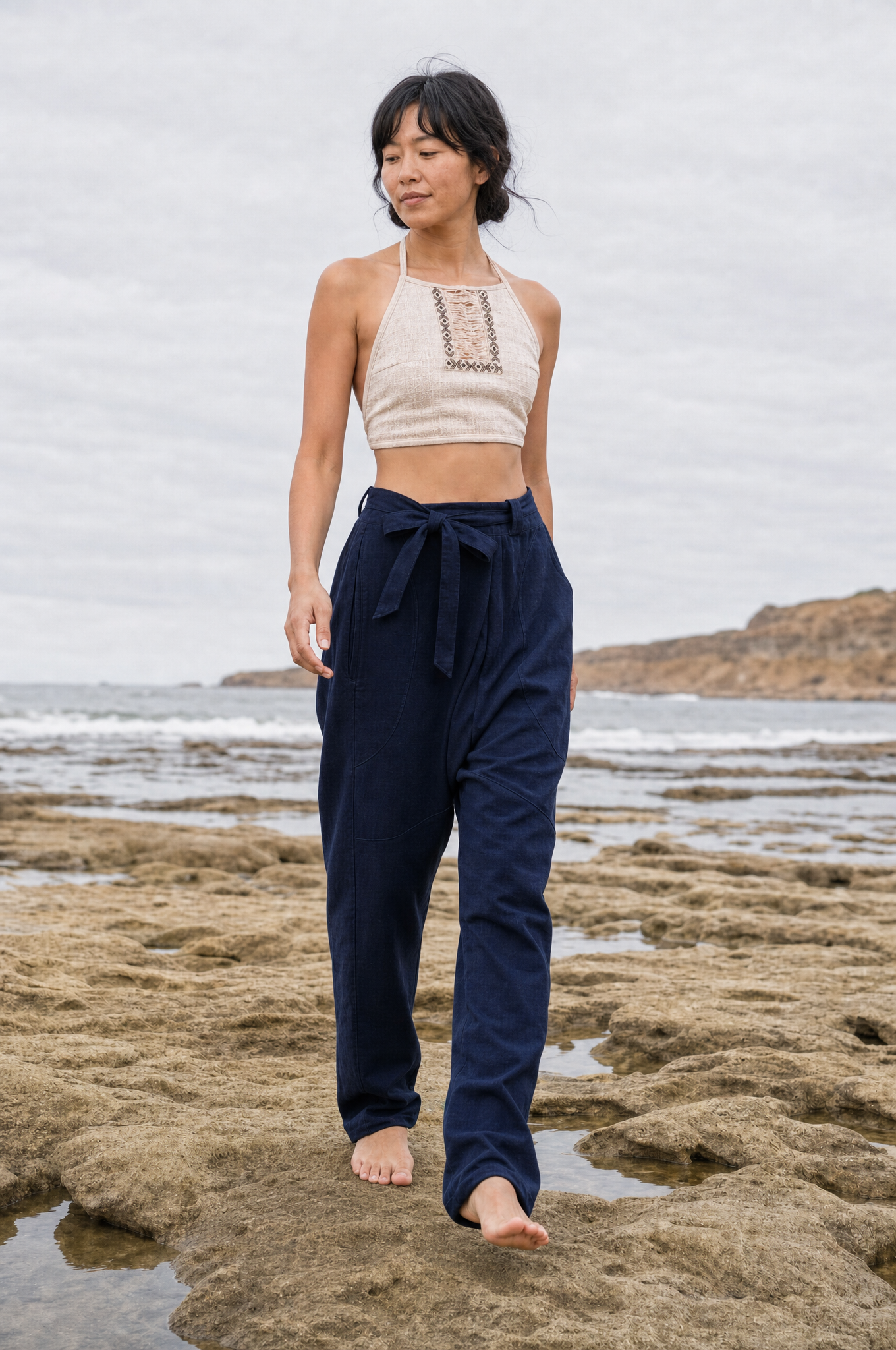 A woman stands barefoot on a rocky beach wearing navy blue drawstring pants and a cream halter crop top with a small embroidered chest detail.