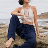 A woman sits barefoot on wet rocks by the ocean, wearing a cream halter crop top with lace-up front detailing and dark navy wide-leg pants.