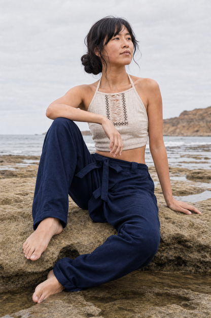 A woman sits barefoot on wet rocks by the ocean, wearing a cream halter crop top with lace-up front detailing and dark navy wide-leg pants.