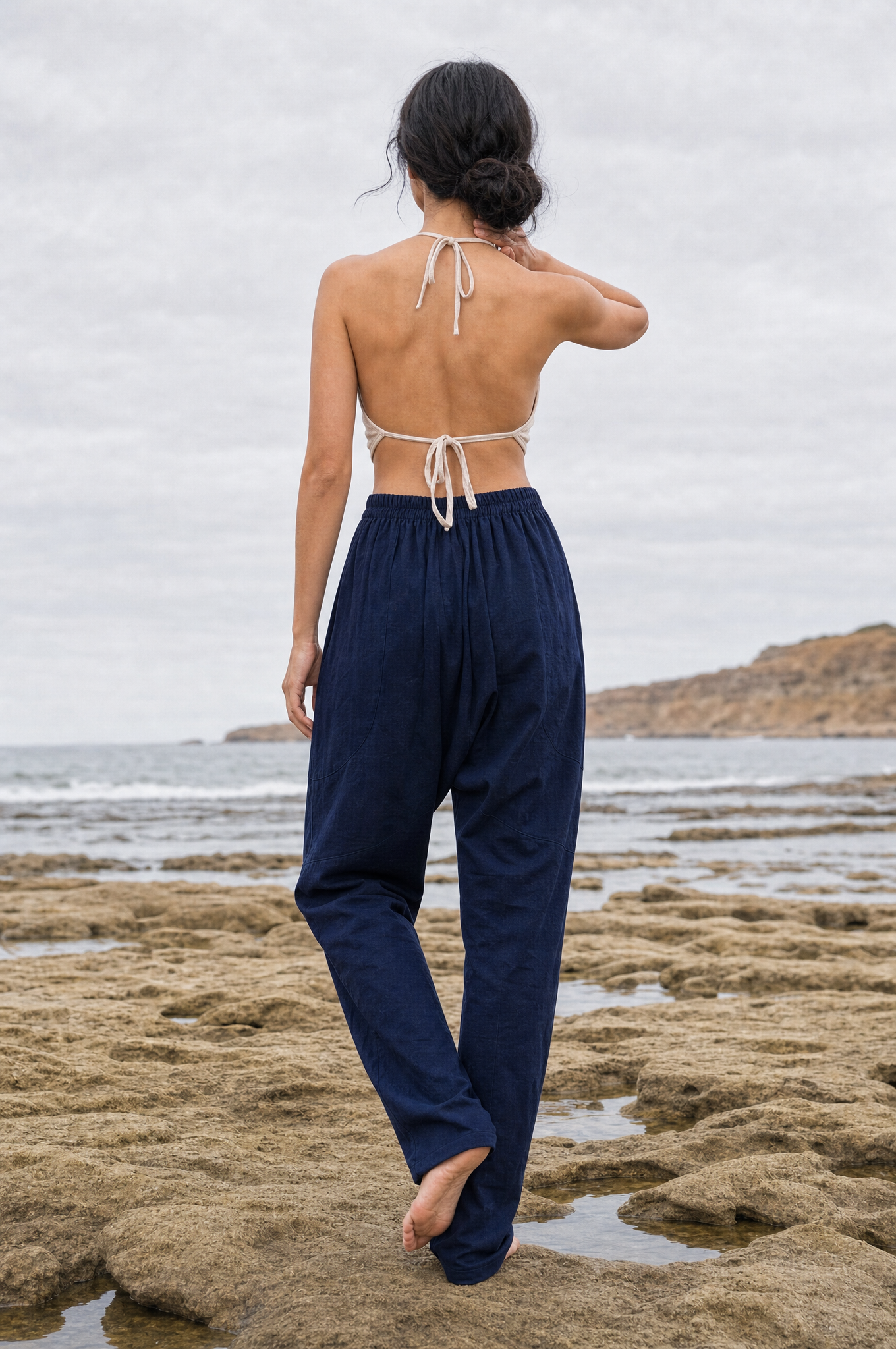 A woman stands on a rocky beach facing the ocean, wearing loose navy pants and a cream halter-style backless top.