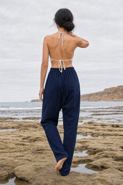 A woman stands on a rocky beach facing the ocean, wearing loose navy pants and a cream halter-style backless top.
