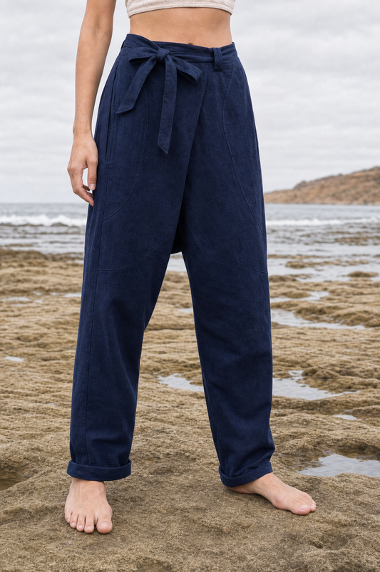A person standing barefoot on a sandy beach wearing loose, high-waisted navy pants with a tie waist and rolled cuffs.