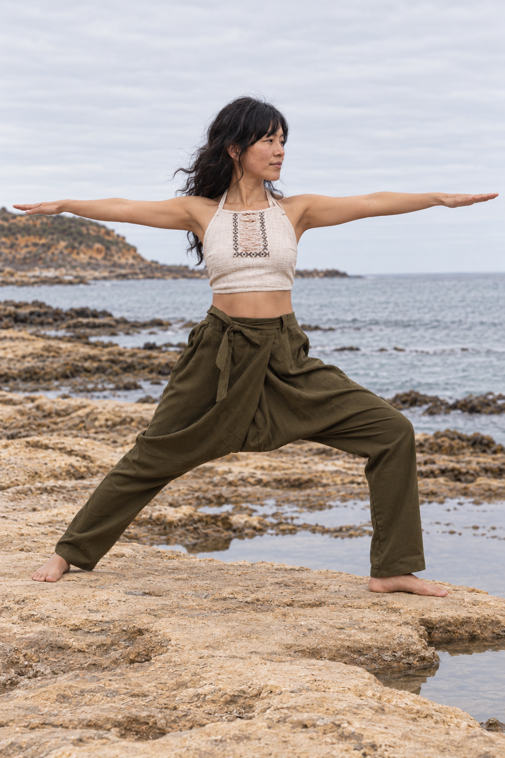 A woman performing a yoga warrior pose on rocky coastal ground with the ocean and cloudy sky in the background. She is wearing an off-white halter crop top and olive green high-waisted loose pants.