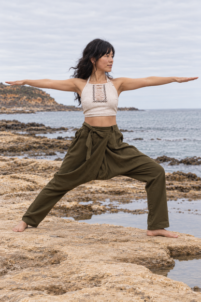 A woman performing a yoga warrior pose on rocky coastal ground with the ocean and cloudy sky in the background. She is wearing an off-white halter crop top and olive green high-waisted loose pants.