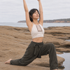A woman practices yoga in a low lunge pose on a rocky beach, wearing a cream halter crop top with embroidery and dark olive green wide-leg pants, with the ocean and cloudy sky in the background.