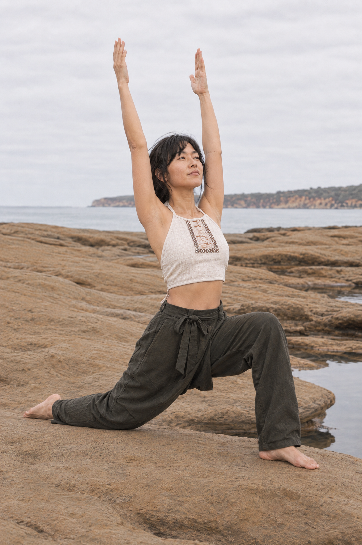 A woman practices yoga in a low lunge pose on a rocky beach, wearing a cream halter crop top with embroidery and dark olive green wide-leg pants, with the ocean and cloudy sky in the background.