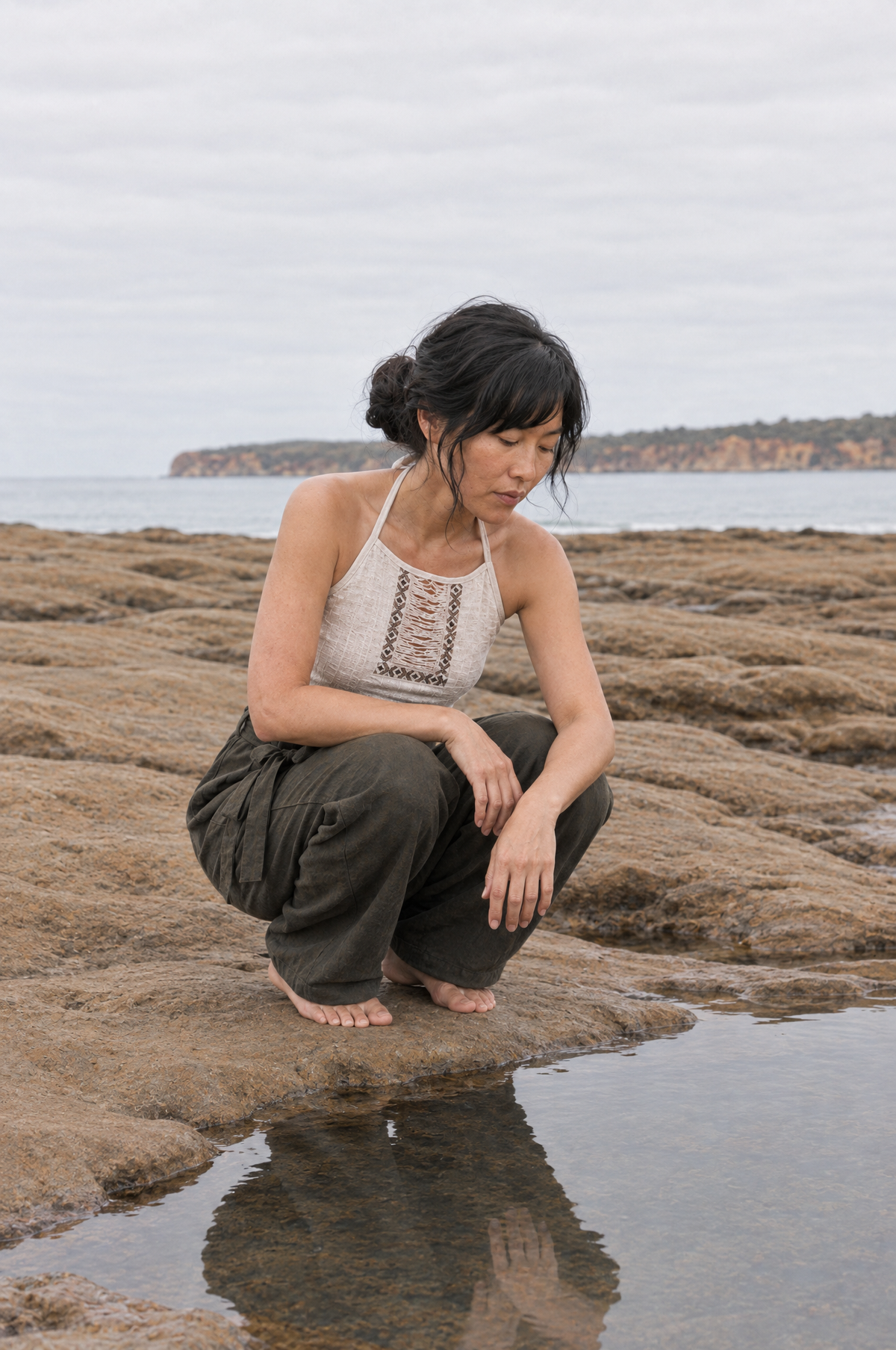 A woman crouches on rocky shoreline ground near a small pool of water, wearing a cream crochet halter top and dark olive cargo pants, with the ocean and distant land visible in the background.