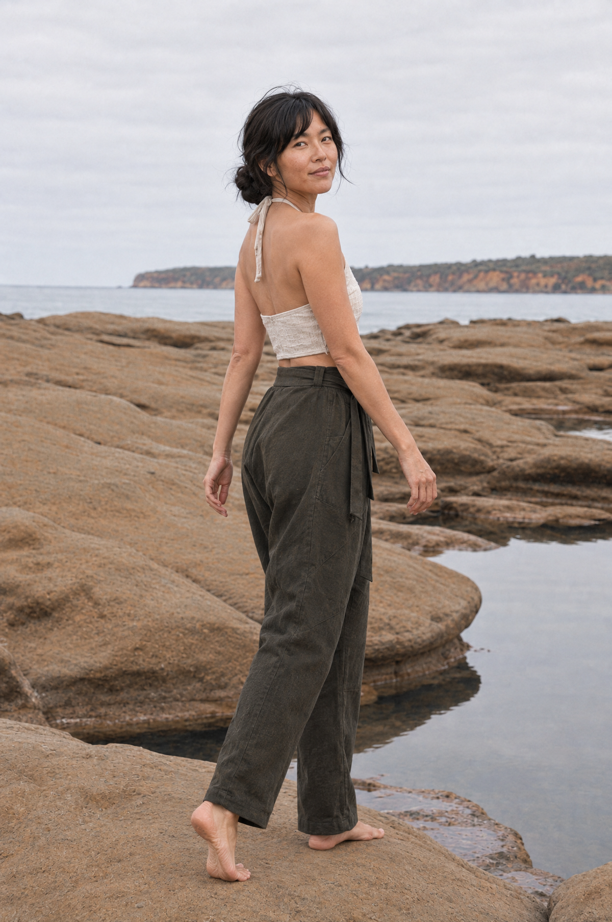 A woman stands barefoot on rocky coastal ground near the water, wearing a light beige halter crop top and dark olive green wide-leg pants.
