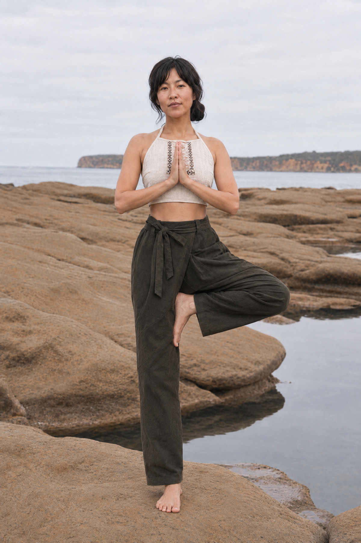 A woman stands in a yoga tree pose on rocky coastal ground, wearing a white halter crop top and dark olive green wide-leg pants. The background shows a calm sea, distant cliffs, and an overcast sky.