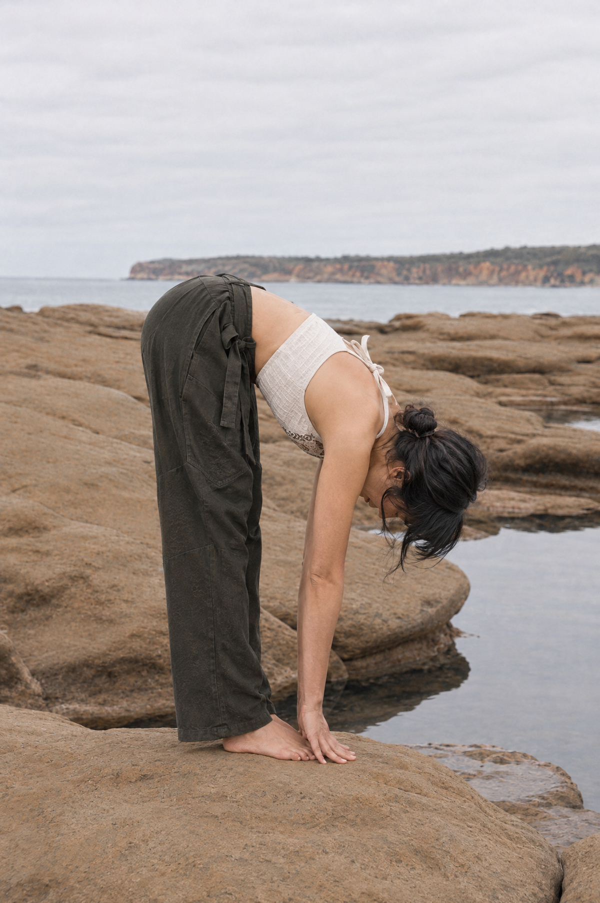 A woman practicing yoga on rocky coastal ground, wearing dark olive green wide-leg pants and a white ribbed tank top, with the ocean and distant cliffs in the background.