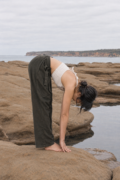 A woman practicing yoga on rocky coastal ground, wearing dark olive green wide-leg pants and a white ribbed tank top, with the ocean and distant cliffs in the background.