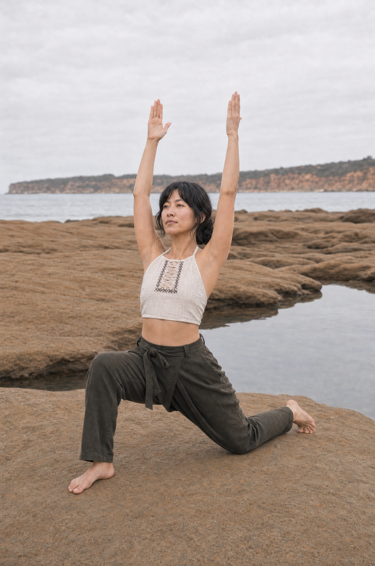 A woman in a yoga pose on a rocky beach with the ocean and distant cliffs in the background, wearing a white crop top and dark olive green pants.