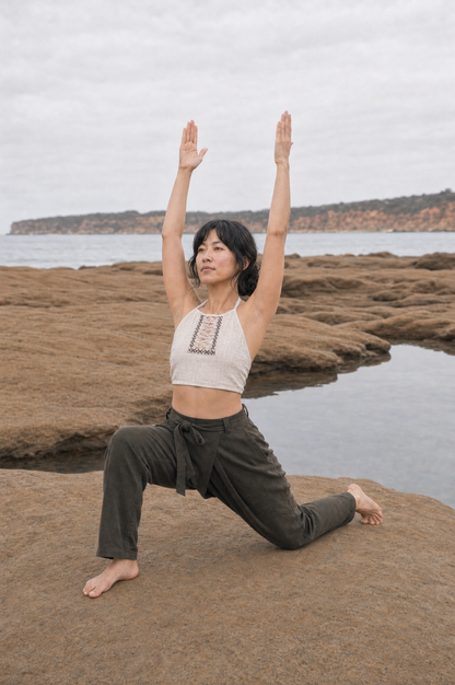A woman in a yoga pose on a rocky beach with the ocean and distant cliffs in the background, wearing a white crop top and dark olive green pants.