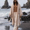 A man walks barefoot on a dark volcanic beach wearing a cream-colored hooded robe and matching drawstring pants with embroidered trim, with ocean waves and rock formations in the background.