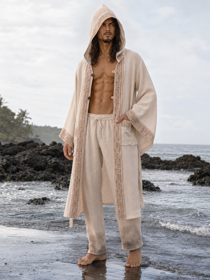 A man stands barefoot on a rocky beach wearing a light beige hooded robe with ornate trim and matching drawstring pants.