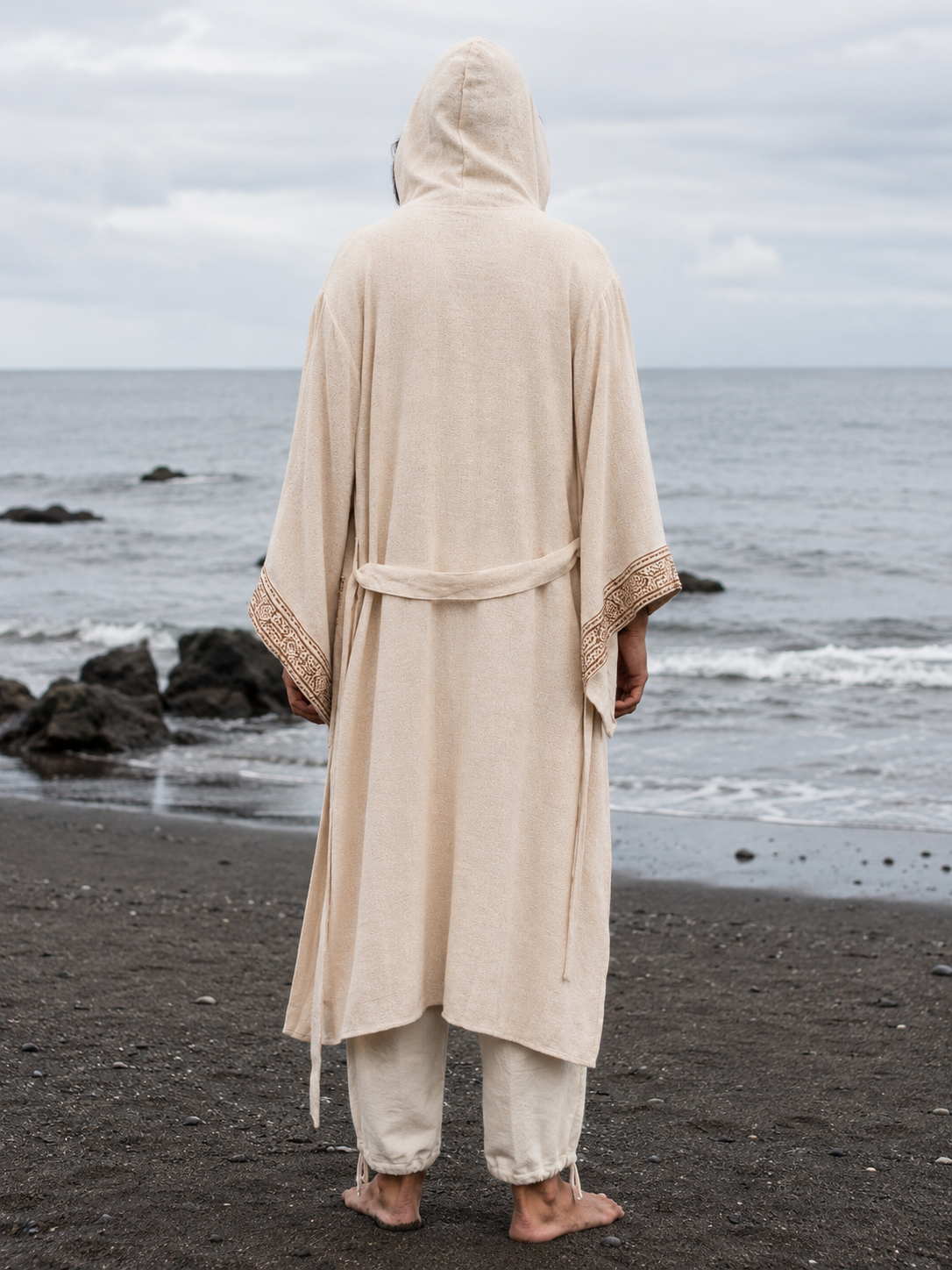 A person standing on a dark beach facing the ocean, wearing a long cream hooded robe with decorative trim on the sleeves and a matching tie belt, paired with light pants.