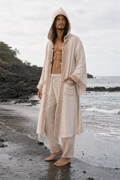 A man stands barefoot on a beach wearing a long cream hooded robe with lace trim and matching loose pants, with ocean and rocky shoreline in the background.