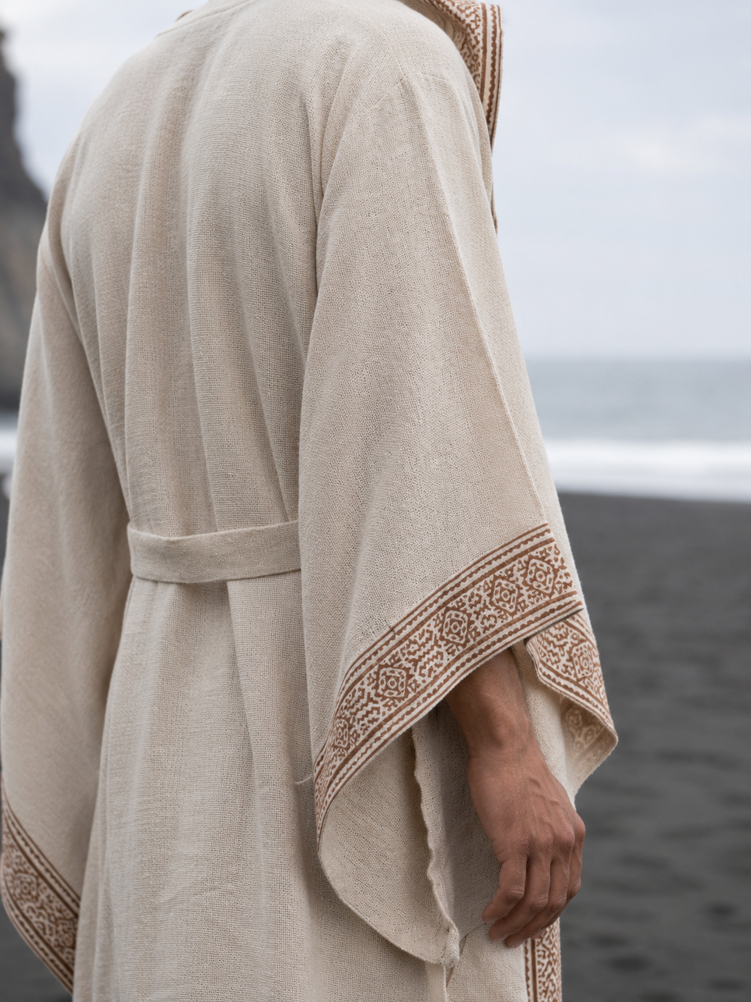 A person wearing a loose beige linen robe with brown patterned trim on the sleeves, standing on a dark beach with the ocean in the background.