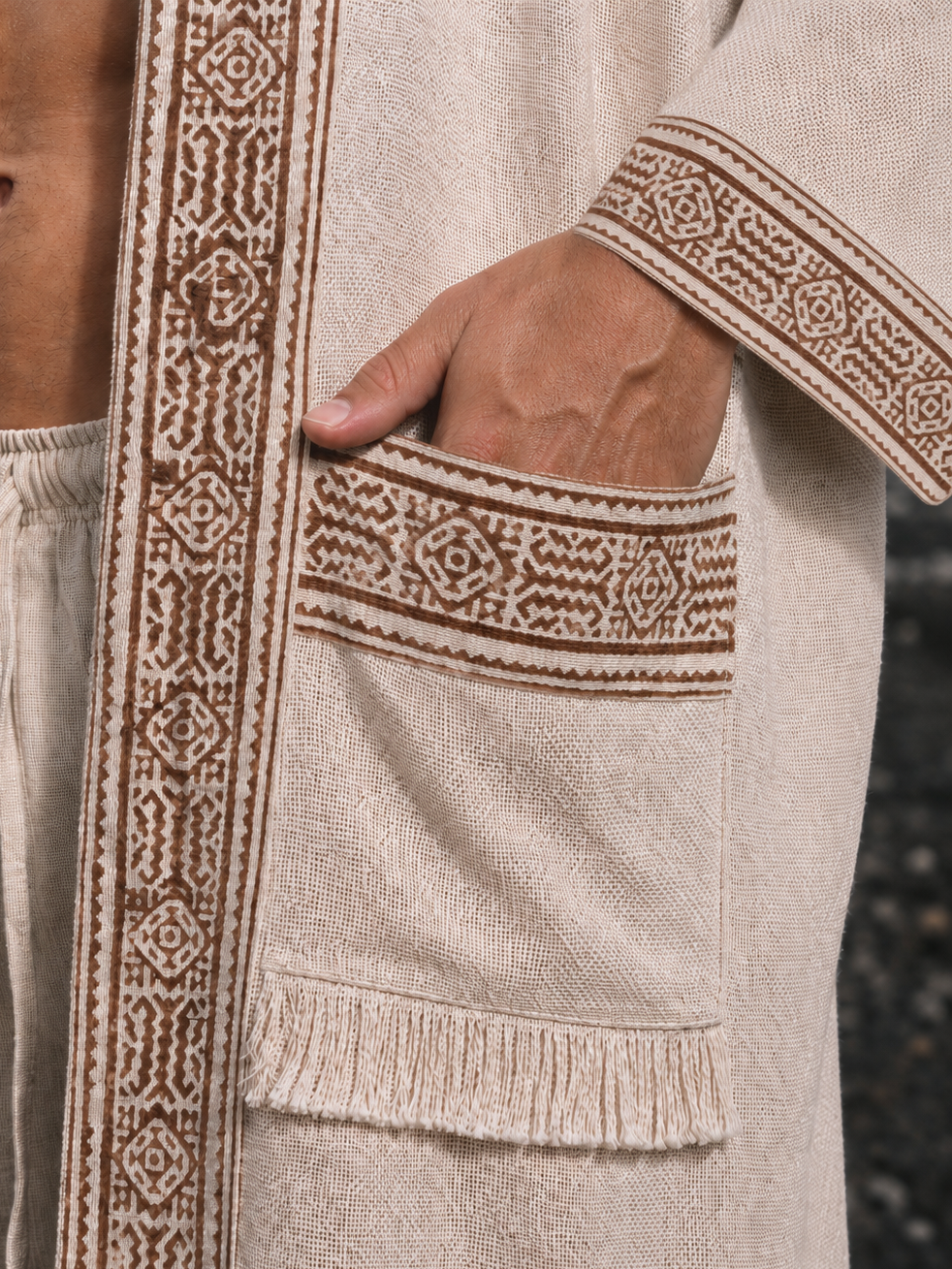 Close-up of a beige woven robe with brown embroidered borders and a large patch pocket, shown on a model with one hand in the pocket.
