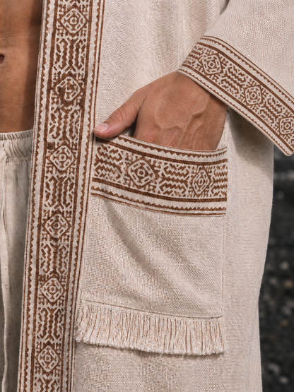Close-up of a beige woven robe with brown embroidered borders and a large patch pocket, shown on a model with one hand in the pocket.
