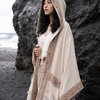 A woman wearing a cream-colored hooded poncho with brown geometric patterns, standing on a black sand beach next to a large dark rock.
