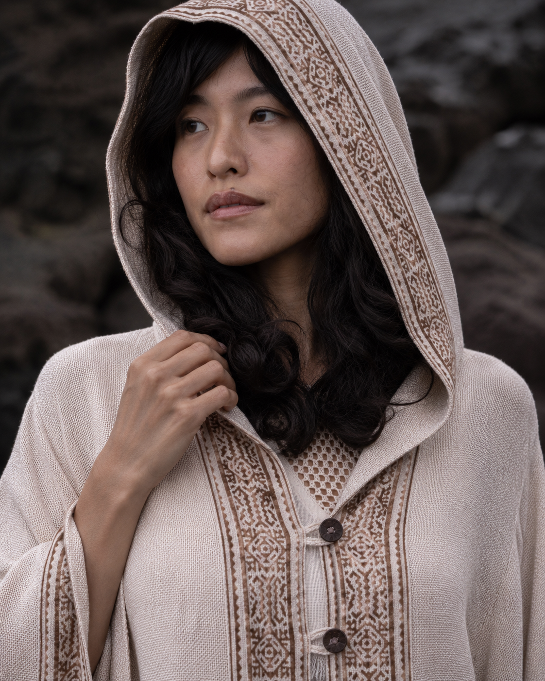 A woman wearing a hooded beige knit poncho with brown embroidered patterns and buttons, set against a dark rocky outdoor background.