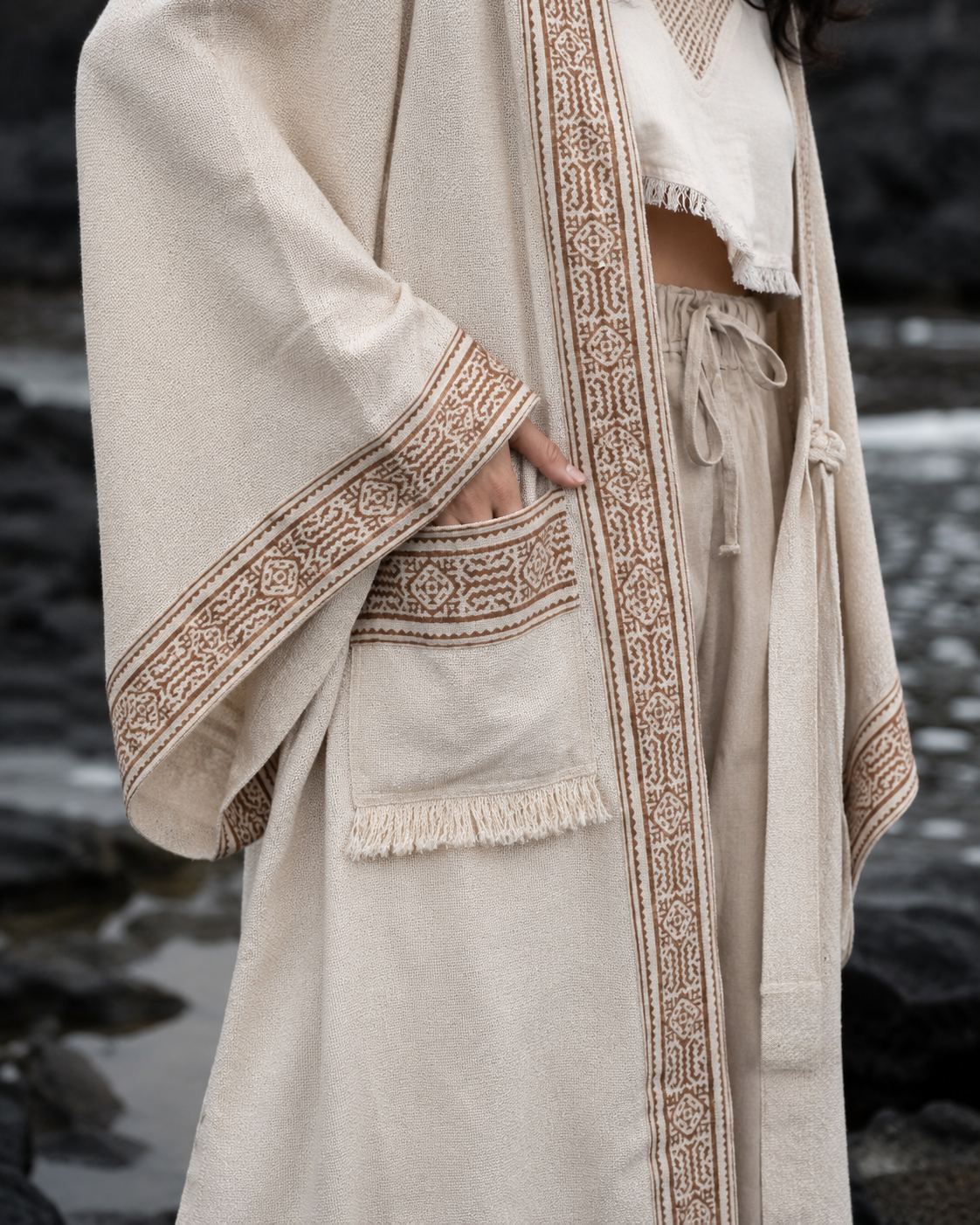 A person wearing a cream-colored linen robe with brown geometric embroidered borders and fringe details, standing outdoors near a rocky shoreline.