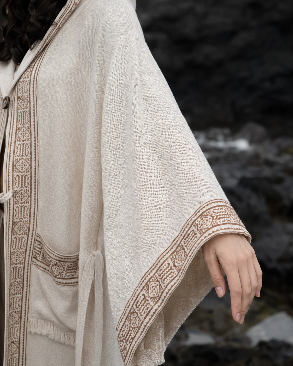 A close-up of a person wearing a cream-colored hooded cloak with brown embroidered borders, set against a dark, rocky outdoor background.