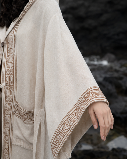 A close-up of a person wearing a cream-colored hooded cloak with brown embroidered borders, set against a dark, rocky outdoor background.