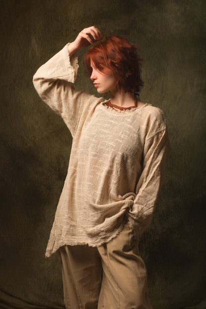 A model with short red hair wearing a loose, textured beige linen tunic top and matching beige trousers, posed against a dark textured studio backdrop.