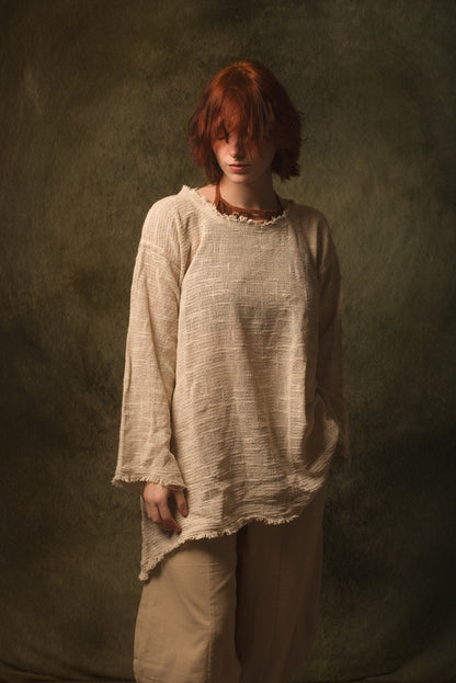 A person with short red hair wearing a loose, textured beige top and matching beige pants, standing against a dark olive-green studio backdrop.