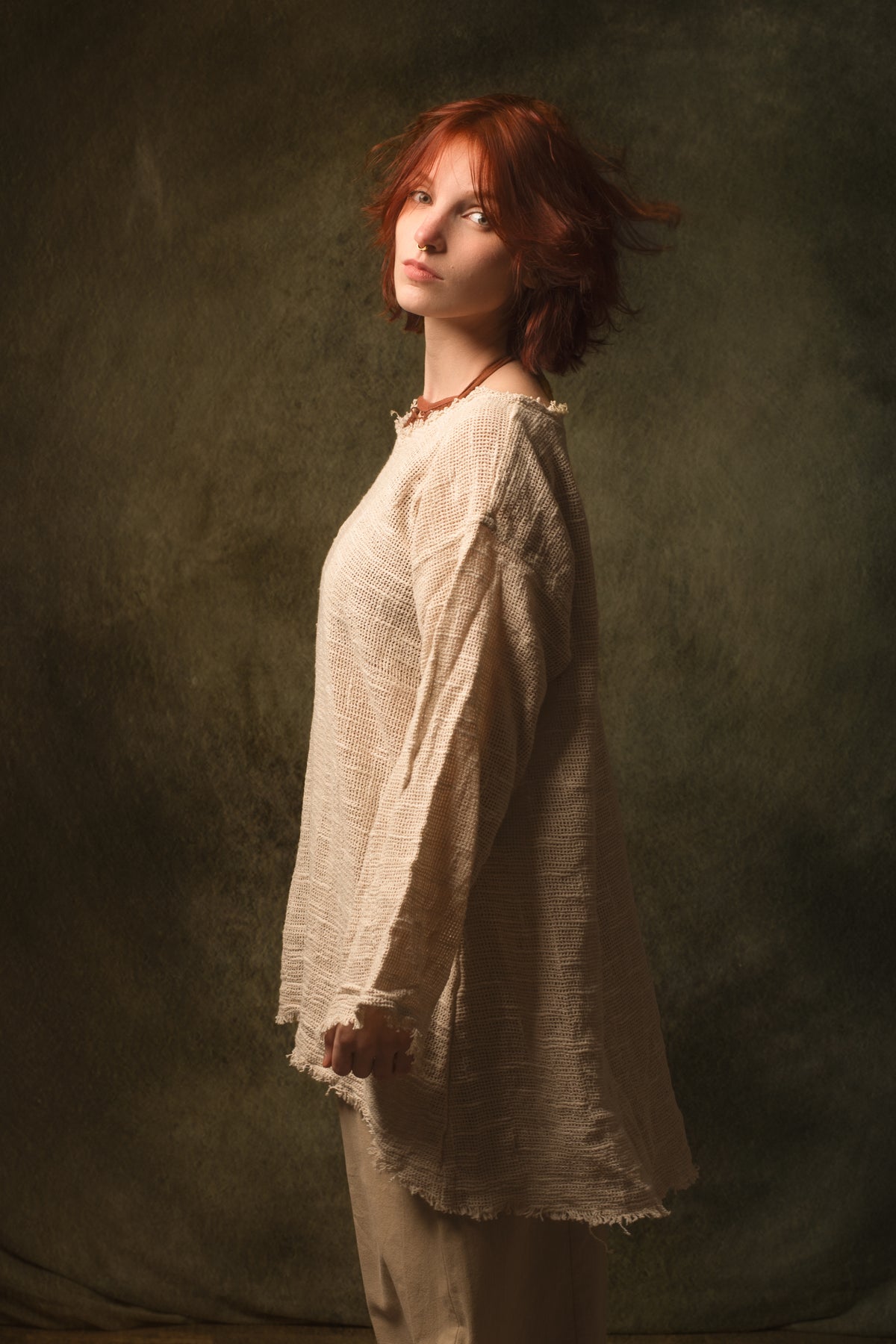A woman with short red hair stands in profile wearing a loose, textured cream linen top and matching pants against a dark olive-green studio backdrop.