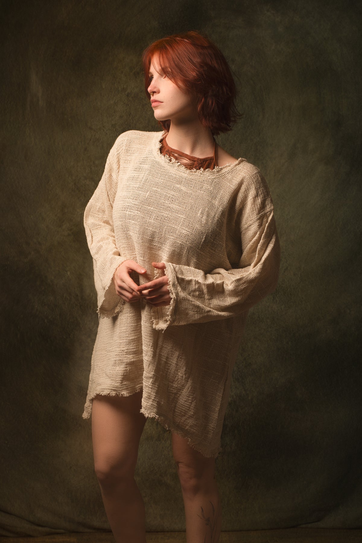 A woman with short red hair wearing a loose, textured cream linen tunic dress with long bell sleeves and a raw, frayed hem, standing against a dark textured studio backdrop.